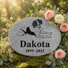 Oval gray memorial garden stone engraved with a black dog illustration and the text 'Dakota 1995-2011', displayed outdoors on moss surrounded by pink and cream roses and greenery.